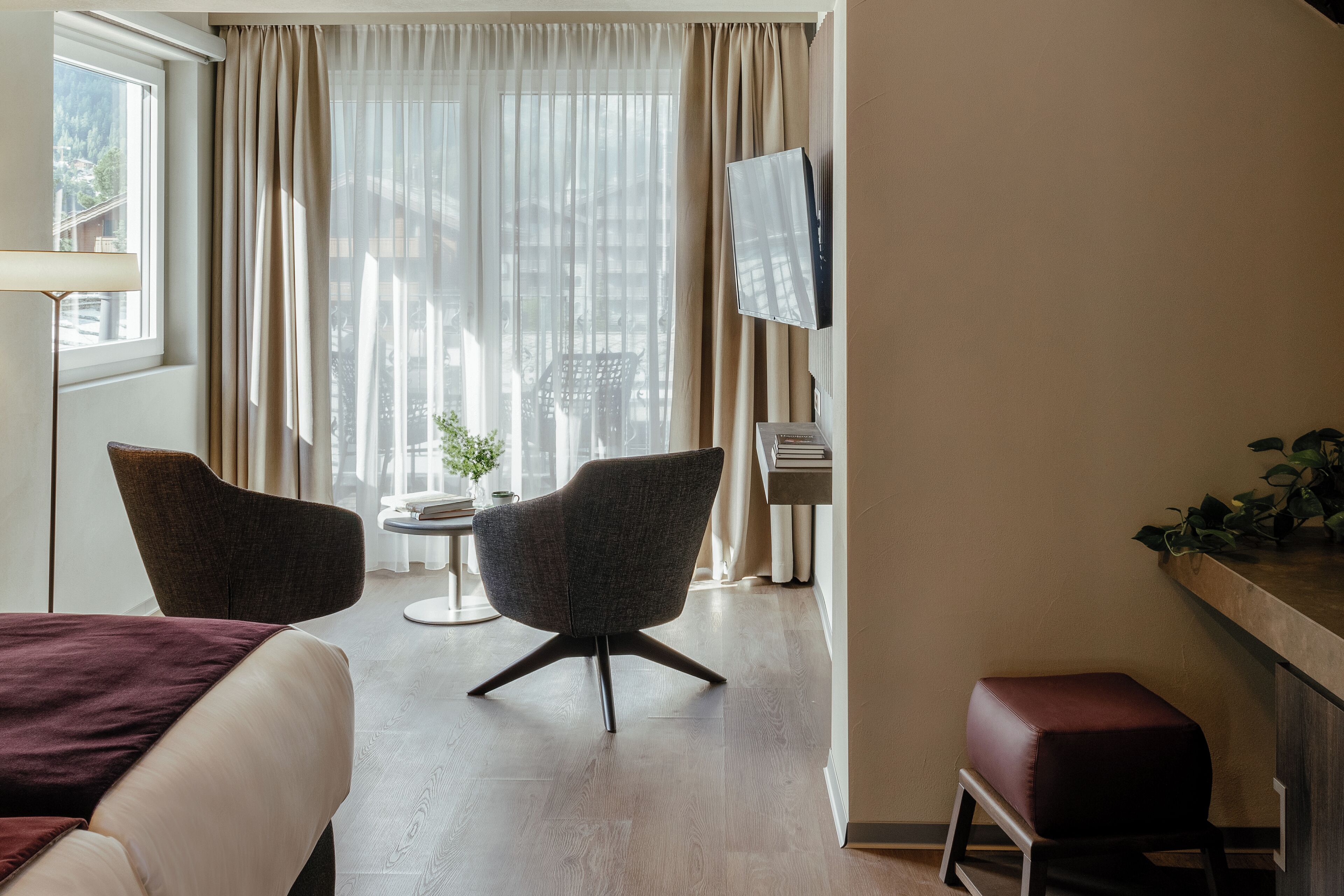 Une chambre d’hôtel avec un lit, deux fauteuils, une petite table, un téléviseur et une fenêtre avec des rideaux.