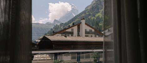 A view of a mountain landscape through a window with curtains, featuring a building with a sloped roof and a modern structure below.
