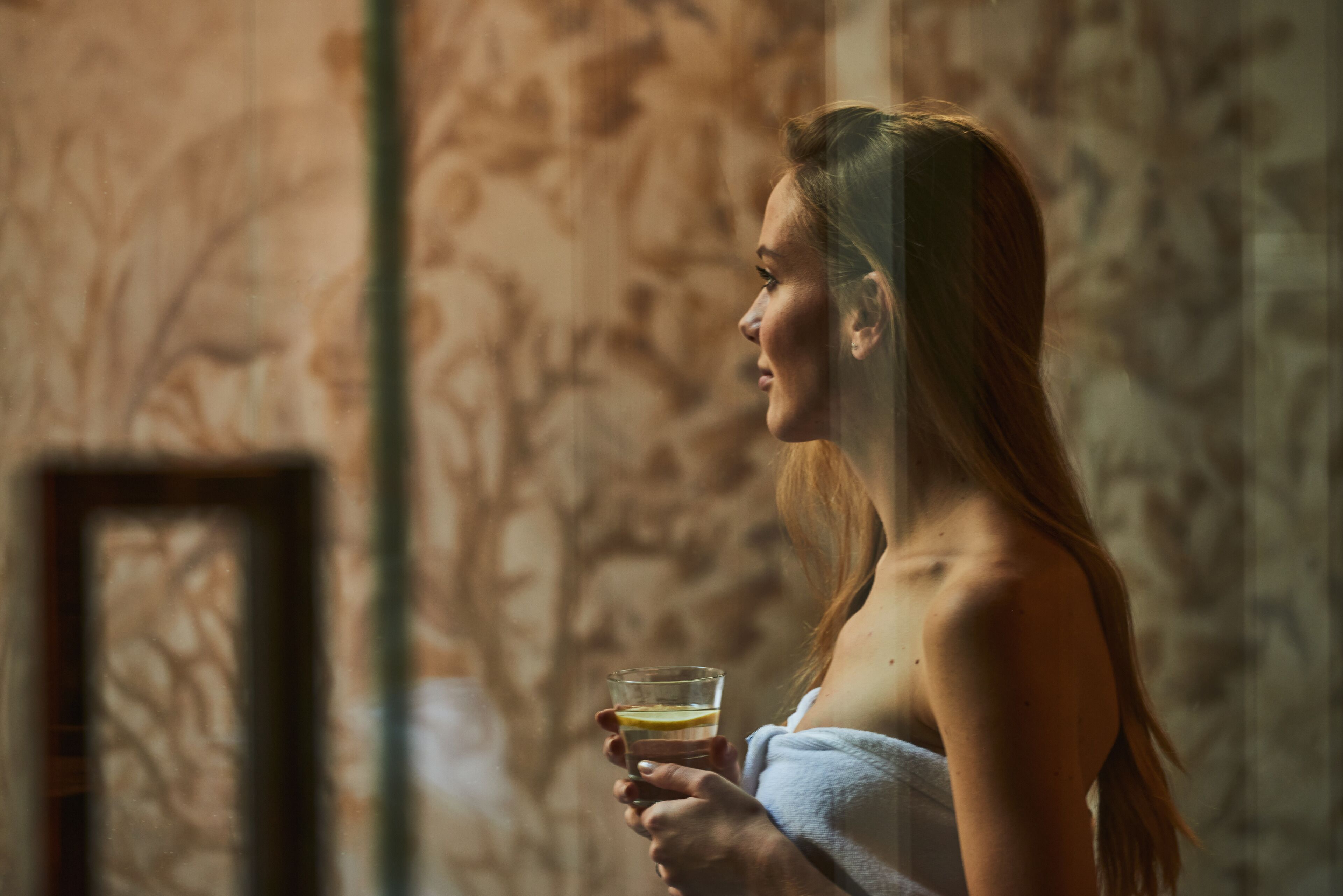 Eine Person hält ein Glas mit einem Getränk und steht vor einer Wand mit Muster.