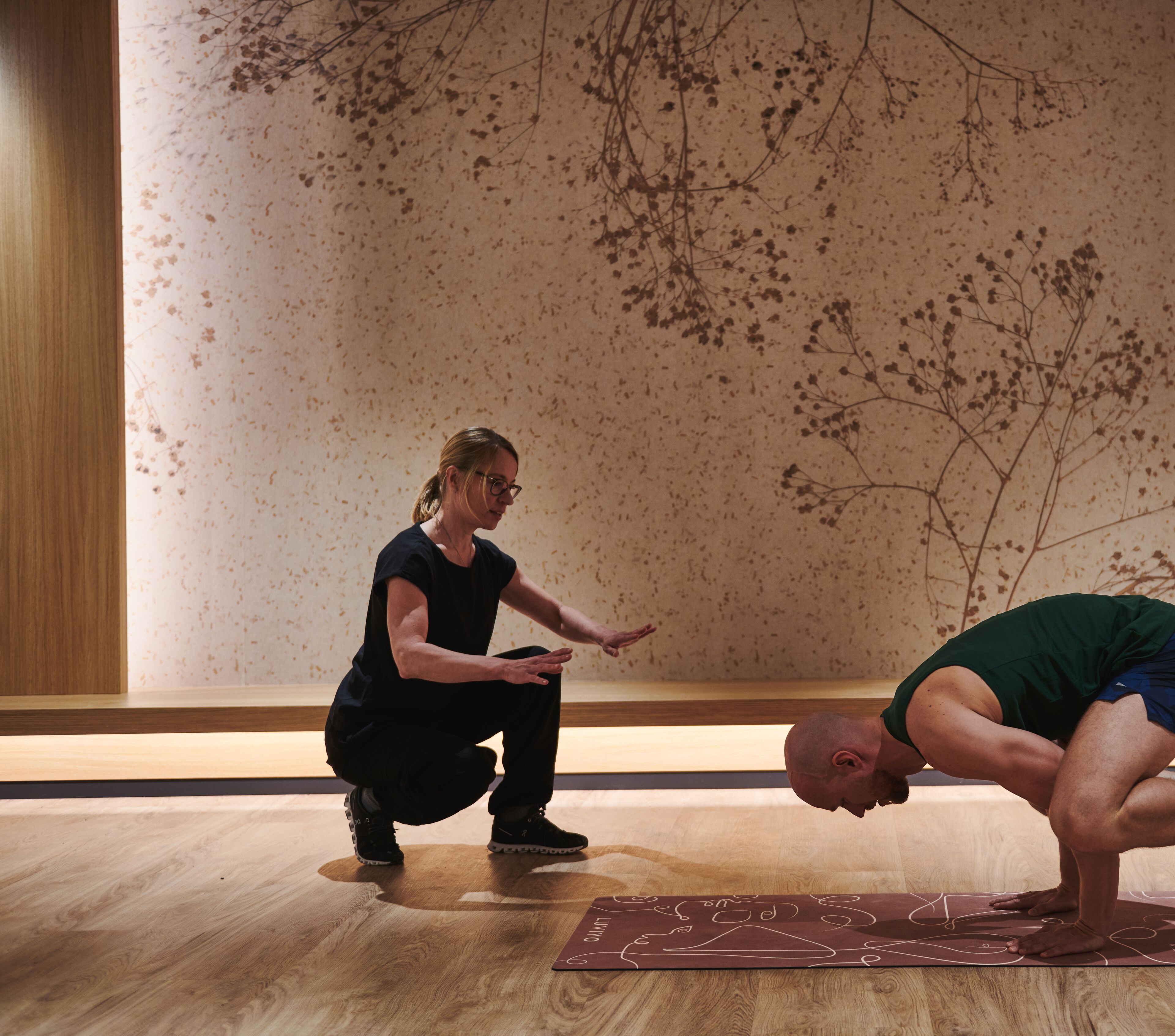 A yoga class in a room with wooden floors and a wall with a tree branch design.