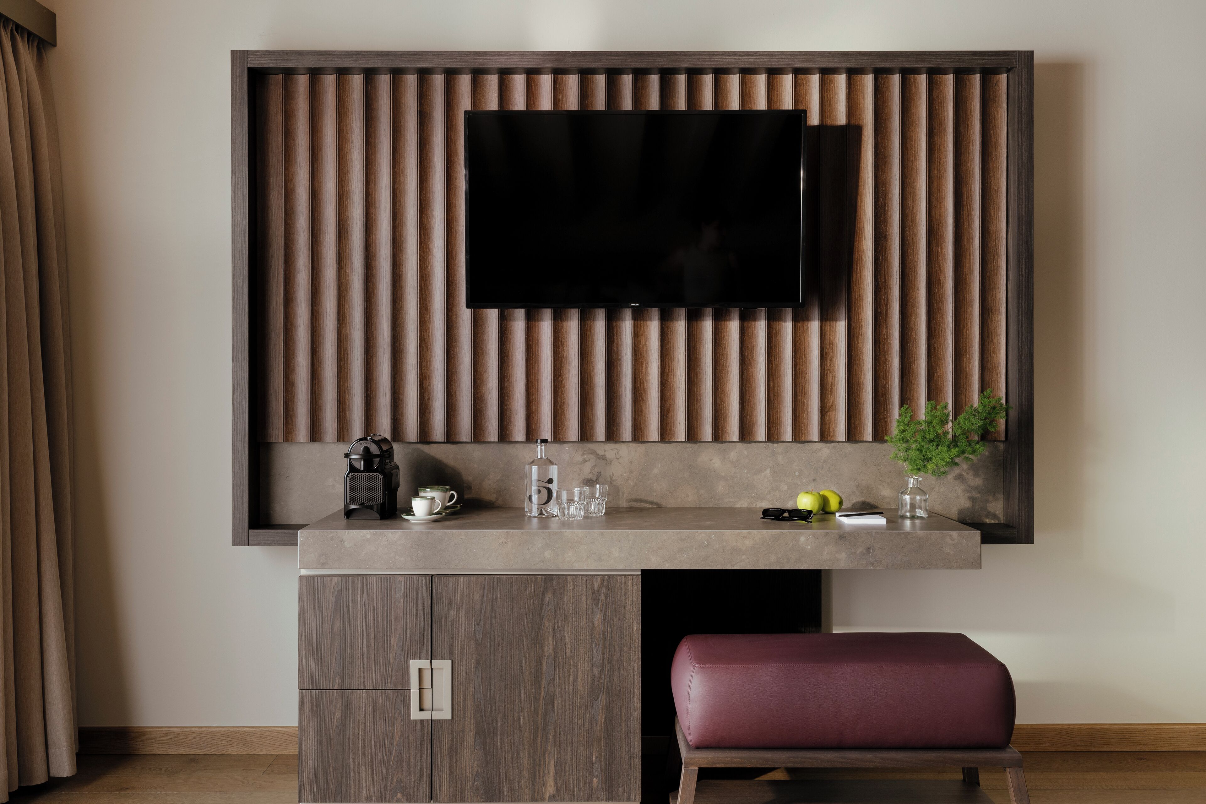 A modern living room with a flat-screen TV mounted on a wooden panel wall, a small coffee table with a kettle, glasses, and a plant, and a red cushioned bench.