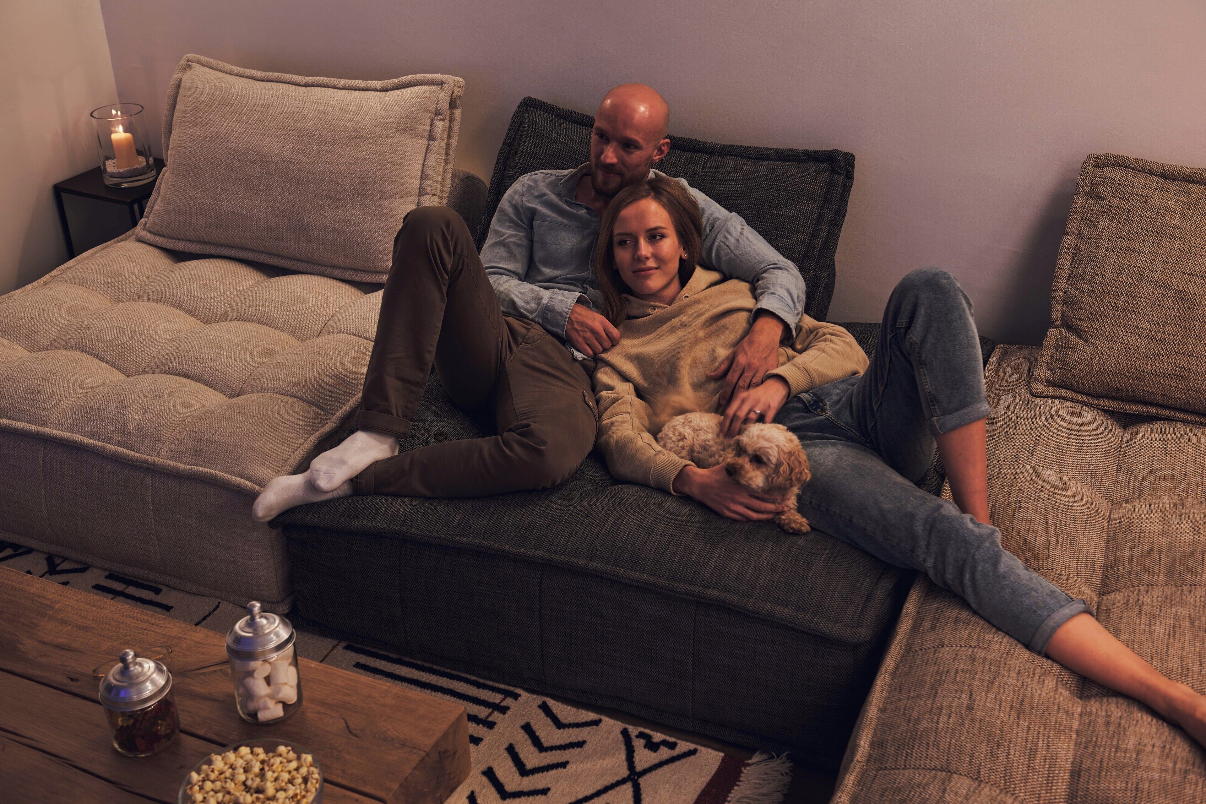 A couple sitting on a sofa with a dog, a candle on a side table, and snacks on a coffee table.