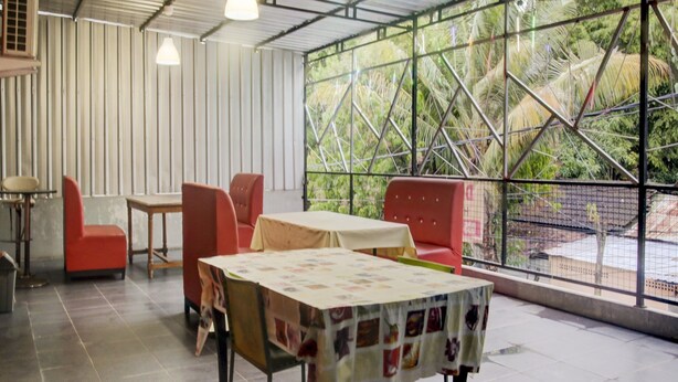 A restaurant with a high ceiling, tiled floor, and a large window with a view of greenery. There are tables and chairs arranged for dining. - SPOT ON 2375 Faris Homestay (Makassar)