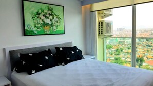 Un dormitorio con una cama grande, una mesita de noche, aire acondicionado instalado en la pared y un amplio ventanal con vistas a la ciudad.