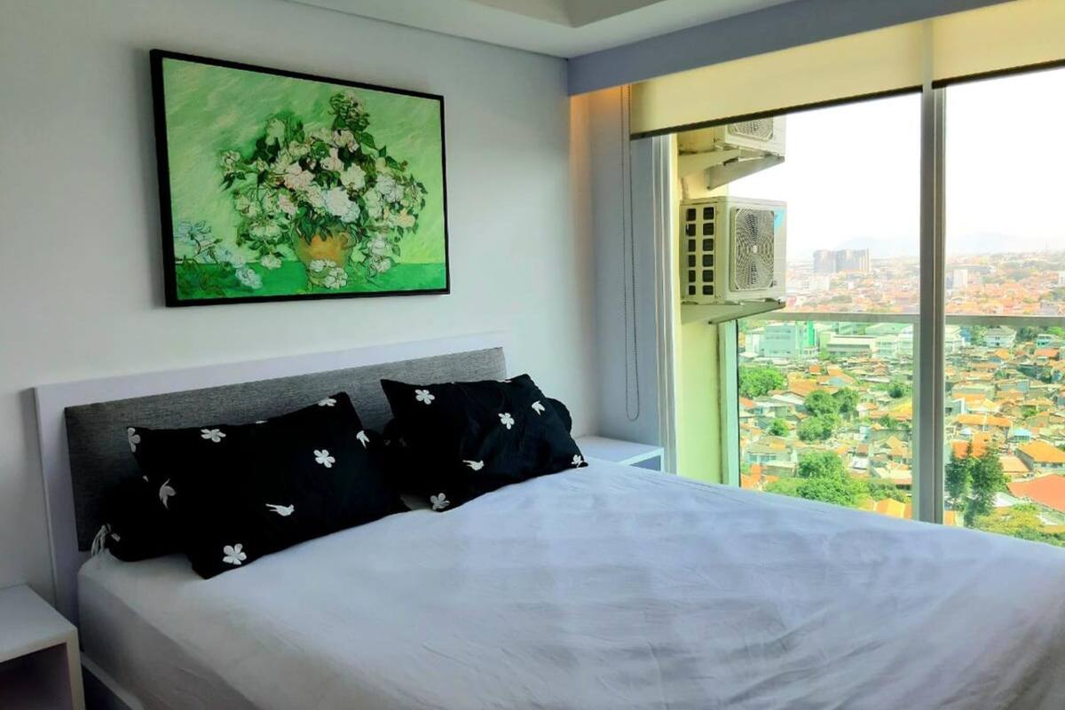 A bedroom with a large bed, a nightstand, a wall-mounted air conditioner, and a large window with a city view.