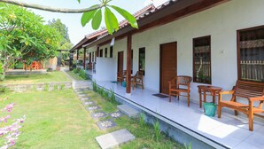 A covered outdoor seating area with wooden benches and tables, a garden with a stone pathway, and a white building with a tiled roof. - Kompyang Rai Guest House (Canggu)