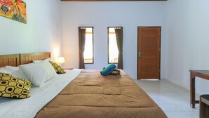 A hotel room with a large bed, two windows with curtains, a wooden door, and a small table. - Kompyang Rai Guest House (Canggu)