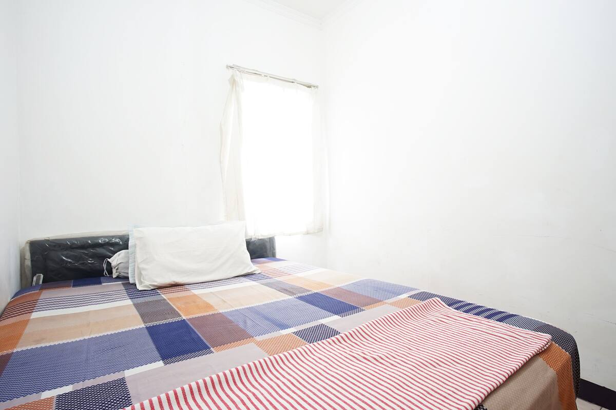 A single bed with a checkered bedspread, a white pillow, and a window with a sheer curtain.