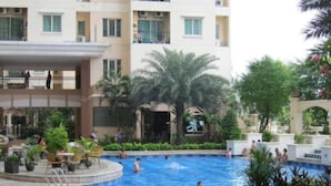 A swimming pool area with a building in the background, surrounded by greenery and palm trees.