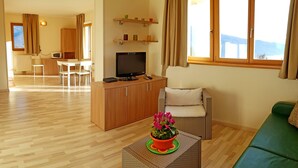 A living room with a television, a green sofa, a wicker coffee table with a flower, and a view of the mountains through the window. - Teresa Appartamento Vista (Tremosine)