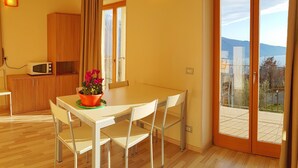 A modern kitchen with wooden cabinets and a microwave, a dining area with a table and chairs, and a sliding glass door leading to an outdoor balcony with a view of the mountains. - Teresa Appartamento Vista (Tremosine)