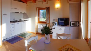 A compact kitchen with white cabinets, a stove, a microwave, and a television. There's a window with curtains, a table with a vase, and a chair. - Teresa Attico Vista (Tremosine)