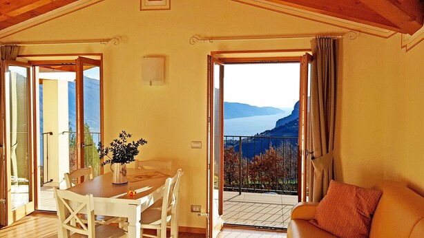 A room with a dining table and chairs, a potted plant, and a sofa. There are open doors leading to a balcony with a view of mountains and a body of water. - Teresa Attico Vista (Tremosine)