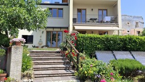A two-story building with a balcony on the upper floor, a stone staircase leading to the entrance, and a garden with various plants and flowers. - Teresa Attico Vista (Tremosine)