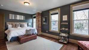 A bedroom with a large bed, a brick accent wall, a window with a view, and a bookshelf with decorative items. - Elegant, King Bed, Privacy,  Free Parking (Washington)