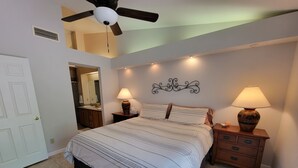Quarto com cama, duas mesas de cabeceira com abajures, ventoinha no teto, luminária na parede e banheiro visível por uma porta aberta.