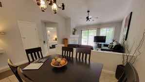 Uma sala de jantar com uma mesa de madeira escura, cadeiras pretas e uma tigela com objetos decorativos. Há um ventilador de teto e uma televisão ao fundo.