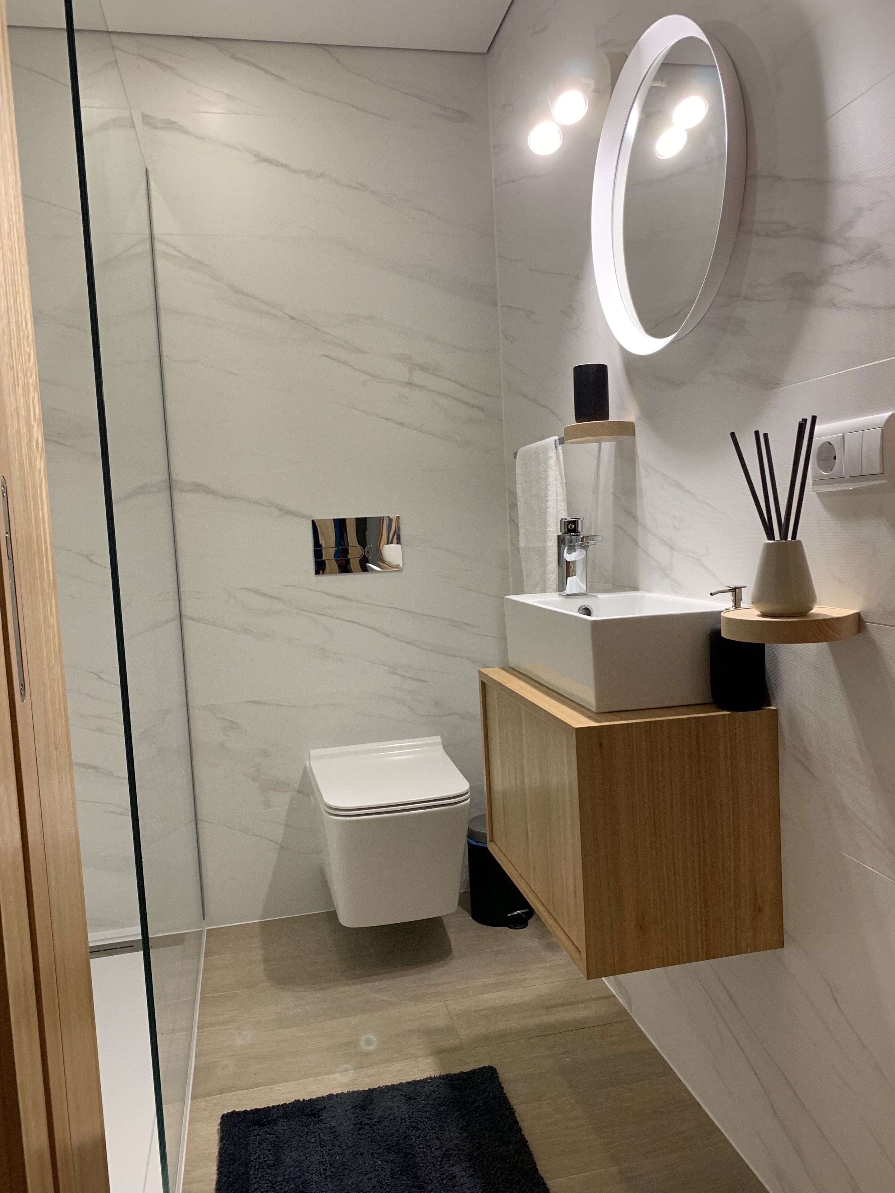 Un baño moderno con inodoro blanco, tocador de madera, espejo y alfombra negra.