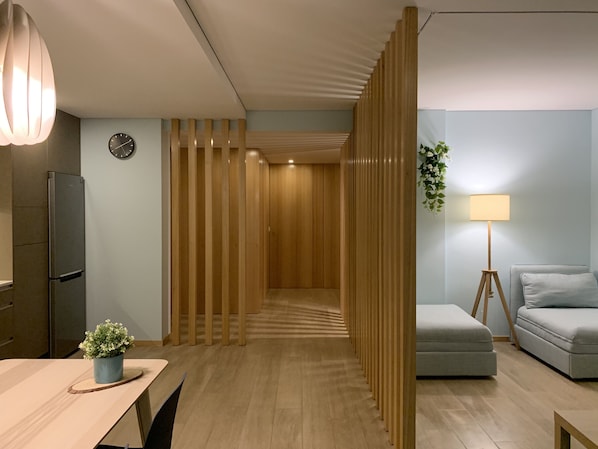 A modern hotel room with a wooden partition, a dining area with a table and chairs, a sofa, a lamp, and a small plant.