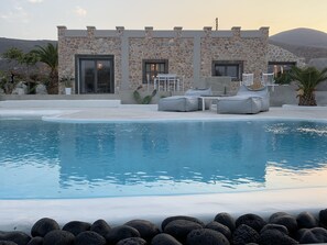 A pool area with lounge chairs and a stone building with large windows. - Villa Beltramo (Santorini)