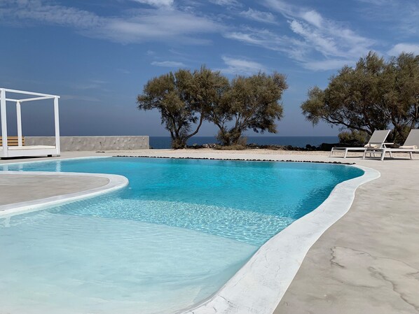 Ein Pool mit weißem Rand, umgeben von Liegestühlen und Bäumen, mit Blick auf das Meer.