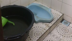 Une salle de bain avec des murs recouverts de carreaux blancs, des toilettes bleues, un lavabo noir et une brosse de nettoyage verte.