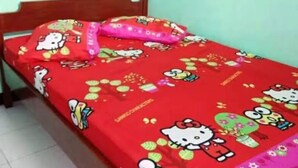 Un lit simple avec une couverture aux motifs Hello Kitty rouge et un oreiller rose, dans une pièce aux murs vert clair et au sol carrelé.