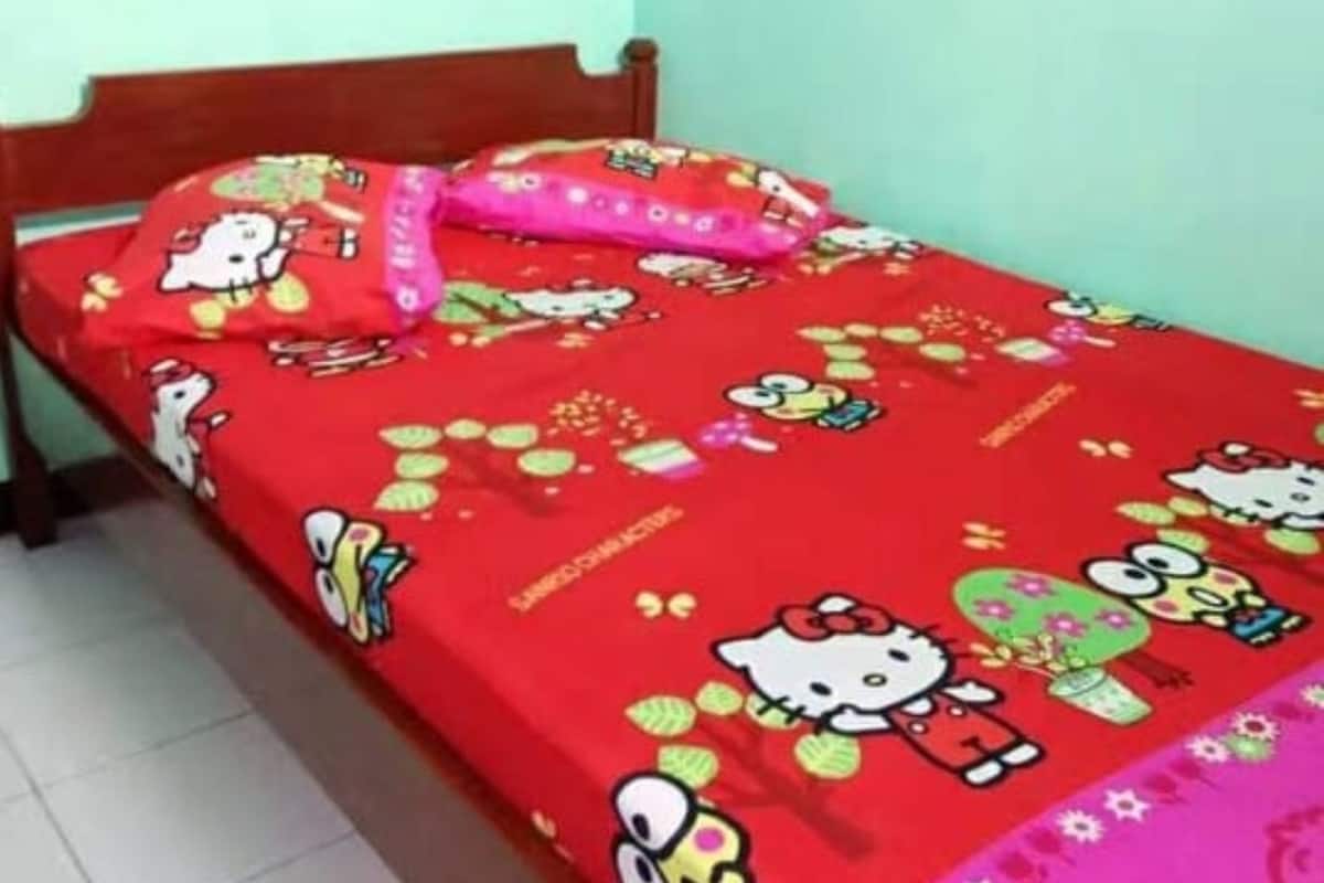A single bed with a red Hello Kitty-themed bedspread and a pink pillow in a room with light green walls and a tiled floor.