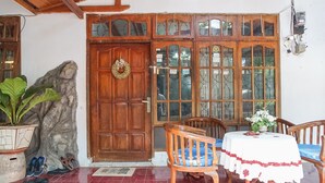 A wooden door with a wreath, a stone wall, a potted plant, and a tiled floor with a table and chairs. - PAS Residence (Manado)