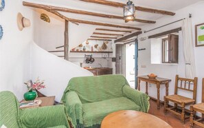 A rustic interior with exposed wooden beams, a green upholstered sofa, a wooden dining table, and a kitchen area with hanging pots.