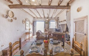 A dining area with a wooden table set for four, surrounded by chairs. There is a basket of fruit on the table, and a bottle of wine. The room has exposed wooden beams on the ceiling and a door with a lattice design.