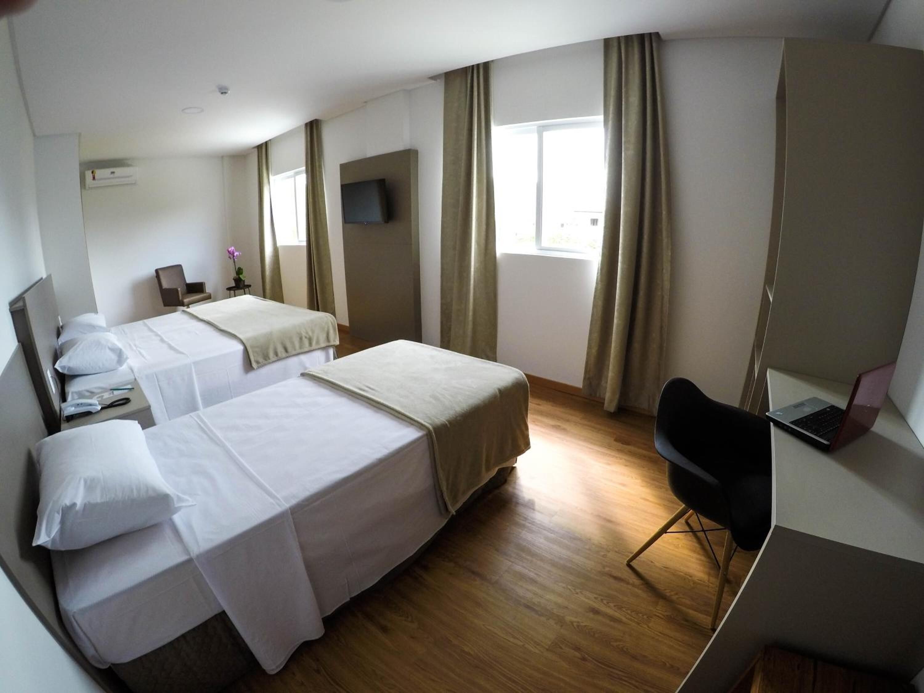 Habitación de hotel con dos camas, un escritorio con una computadora portátil, una silla y un televisor en la pared.