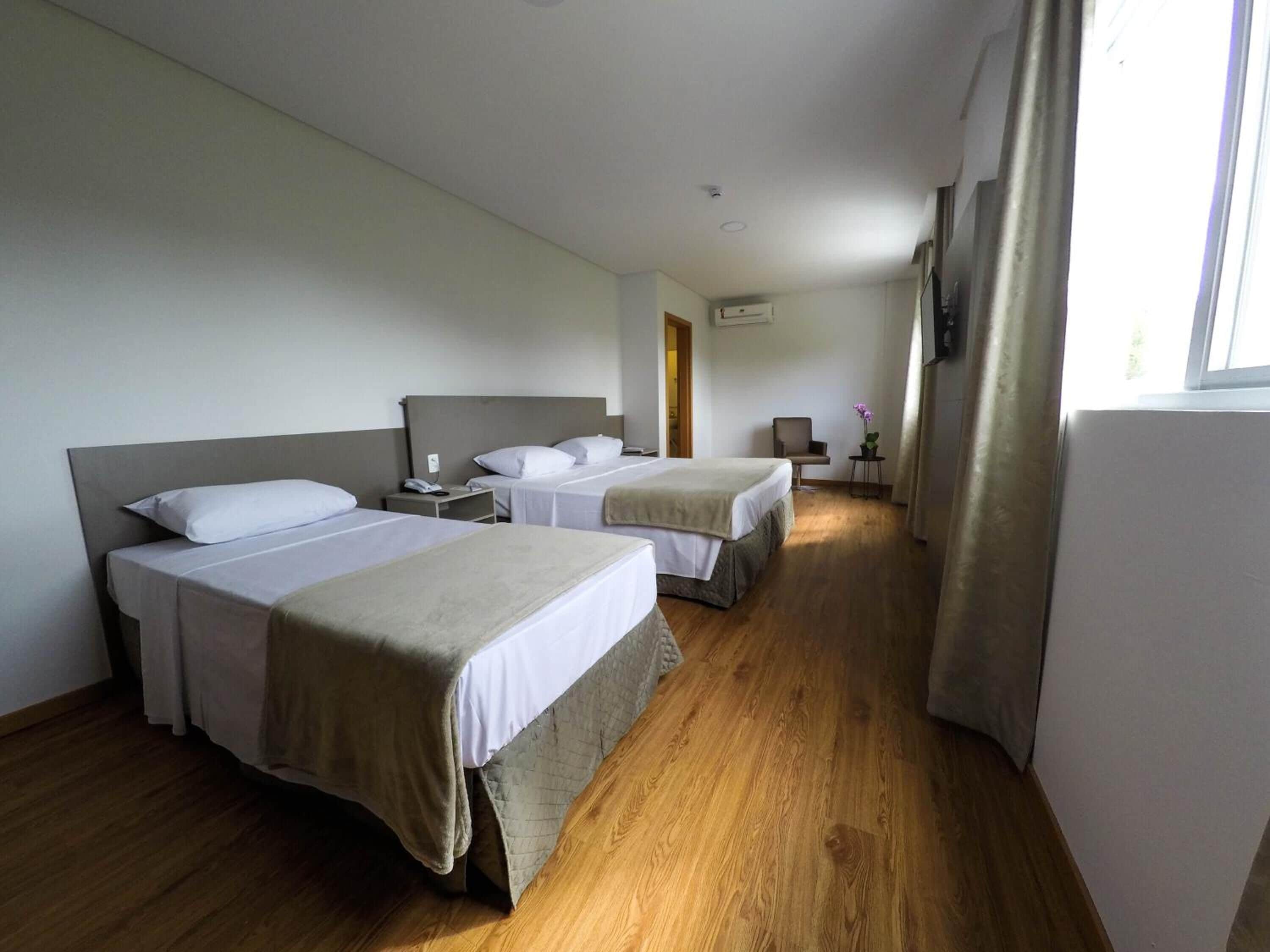 A hotel room with two single beds, a small table, a chair, and a window with curtains.