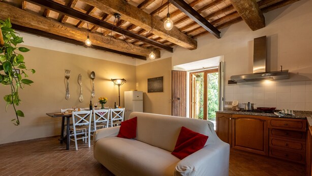 A cozy living room with a white sofa, red cushions, a dining table with chairs, and a kitchen area with wooden cabinets and a built-in stove. - Agritourism San Carlo - apartment Trilo1 (Pomarance)