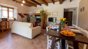 A cozy living room with a white sofa, wooden ceiling beams, a fireplace, and a dining area with a table set for two. - Agritourism San Carlo - apartment Trilo1 (Pomarance)
