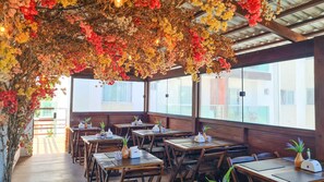 An indoor dining area with wooden tables and chairs, a ceiling adorned with hanging flowers, and large windows providing a view of the outside. - Porto Sol Flats by AFT (Ipojuca)