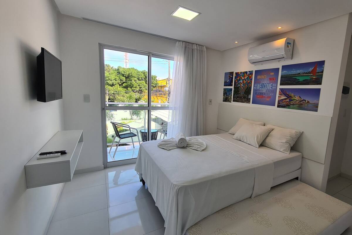 Un dormitorio moderno con una cama grande, una mesita de noche pequeña, un televisor en la pared y un balcón con vistas a zonas verdes y edificios.