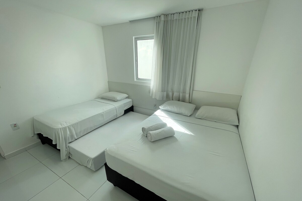 A small, clean room with two single beds, a window with sheer curtains, and a tiled floor.