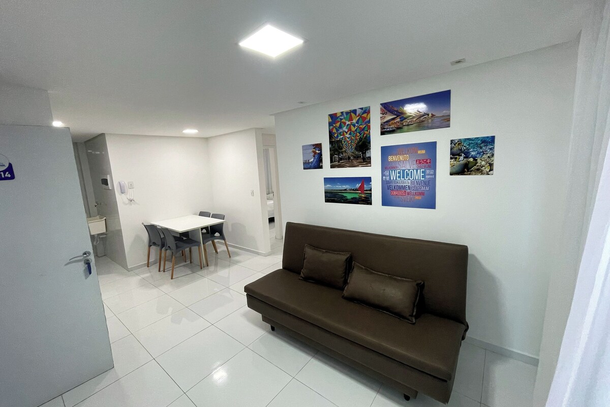 A modern living room with a brown sofa, a dining table with chairs, and a kitchen area in the background.