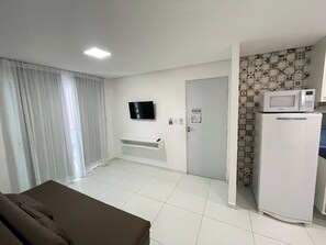 A room with a flat-screen TV mounted on the wall, a white refrigerator with a microwave on top, a door with a sign, and a window with curtains. - Porto Sol Flats by AFT (Ipojuca)
