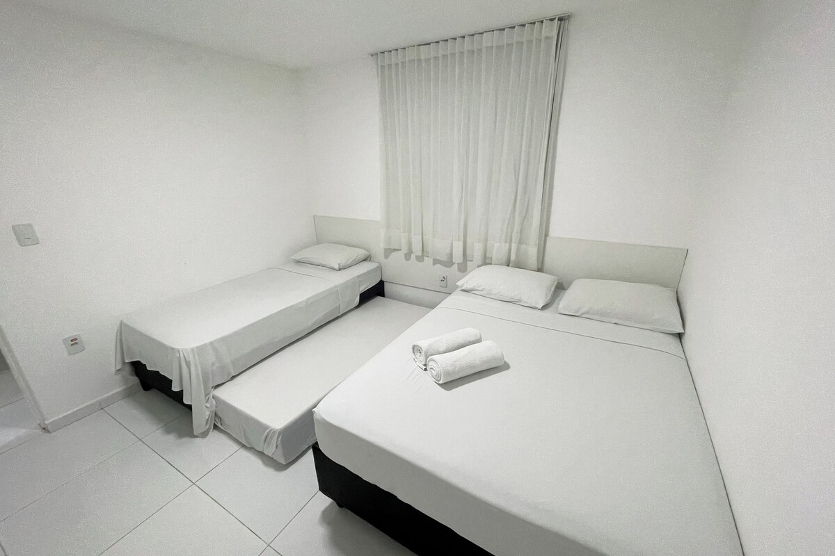 A room with two beds, a window with curtains, and a white tiled floor.