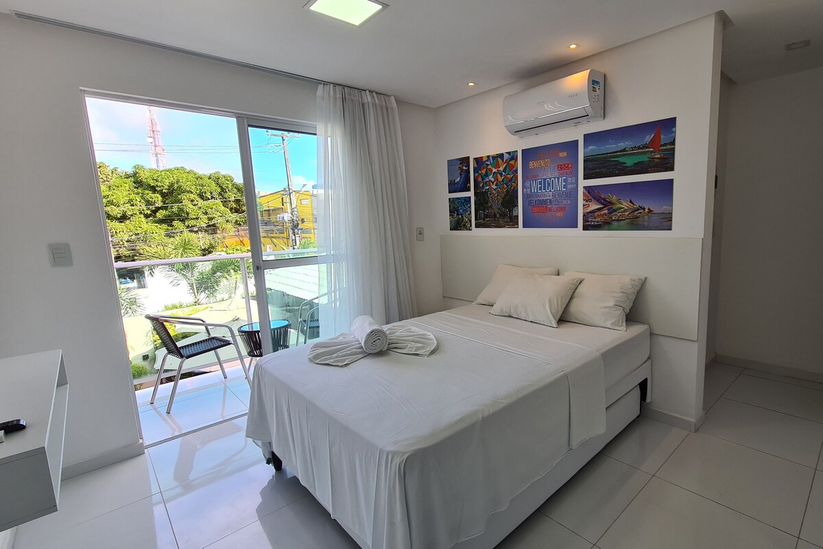 A modern hotel room with a large bed, a balcony with a chair, and a view of greenery outside.