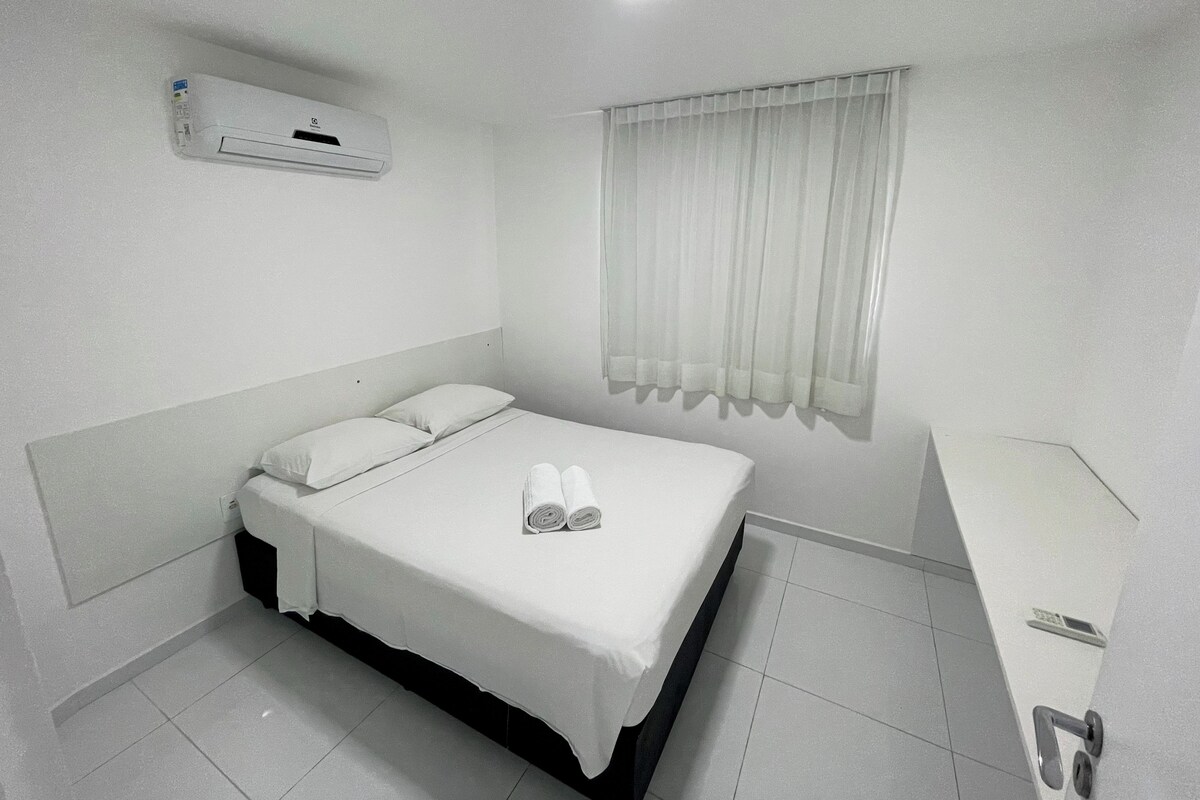 A hotel room with a bed, a window with curtains, a bedside table, and an air conditioning unit.