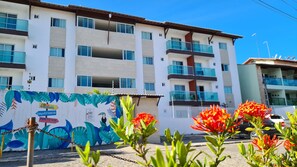 A multi-story hotel with balconies, a mural with tropical leaves and a toucan, and a chain-link fence in the foreground. - Porto Sol Flats by AFT (Ipojuca)