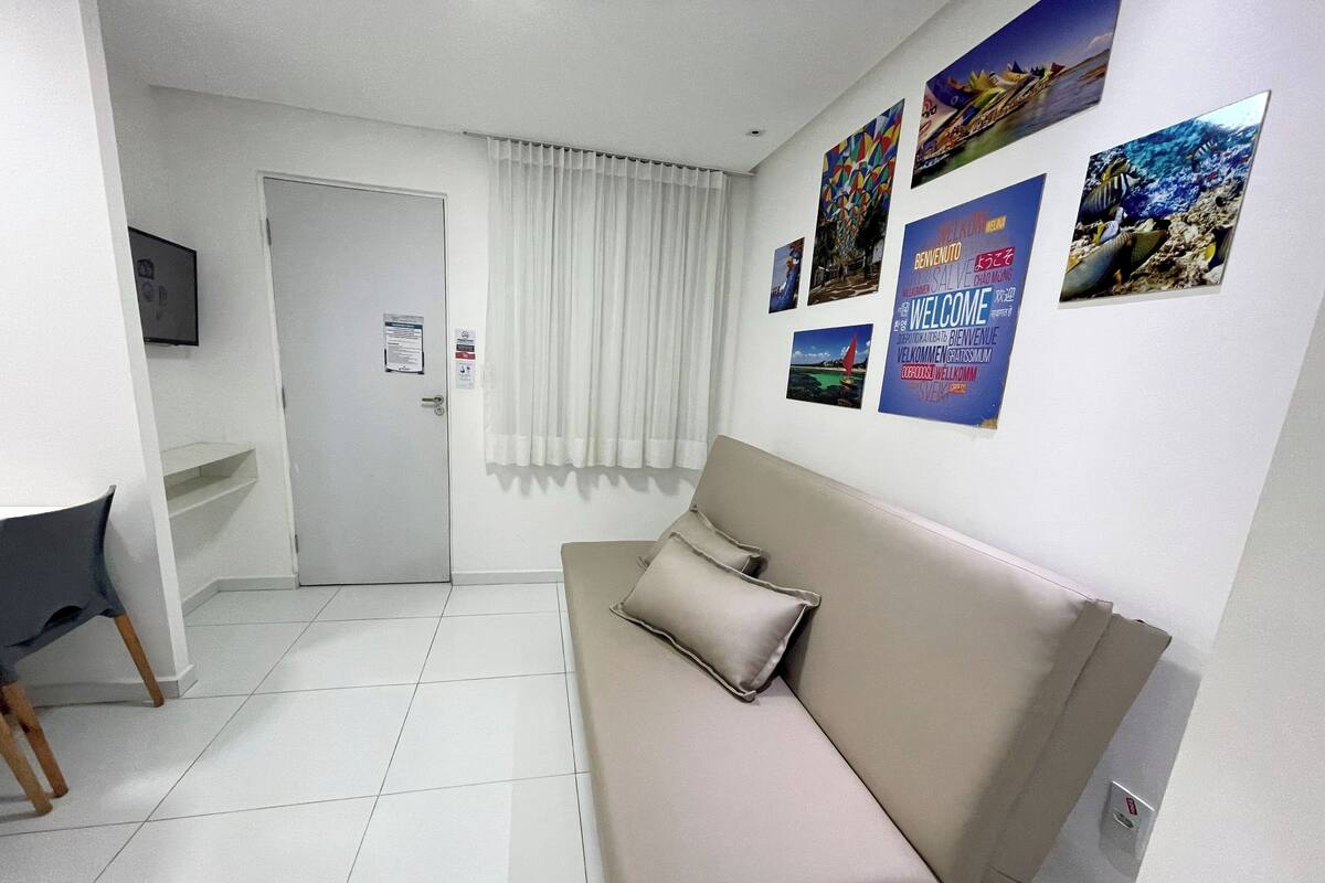 Una habitación con sofá color beige, un escritorio pequeño y un televisor en la pared. Hay varios carteles en la pared, y una puerta con un letrero.