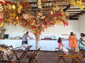 A cafe interior with wooden tables and chairs, a counter with a coffee machine, and a decorative plant arrangement. - Porto Sol Flats by AFT (Ipojuca)