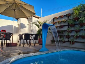Una piscina con una fuente azul, mobiliario exterior y plantas en macetas.