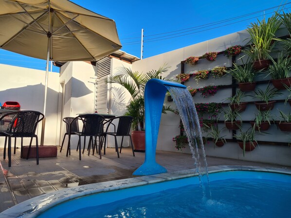 Una piscina con una fuente azul, mobiliario exterior y plantas en macetas.