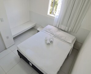 A single-bed room with a white bed, a small bedside table, and a window with curtains. - Porto Sol Flats by AFT (Ipojuca)