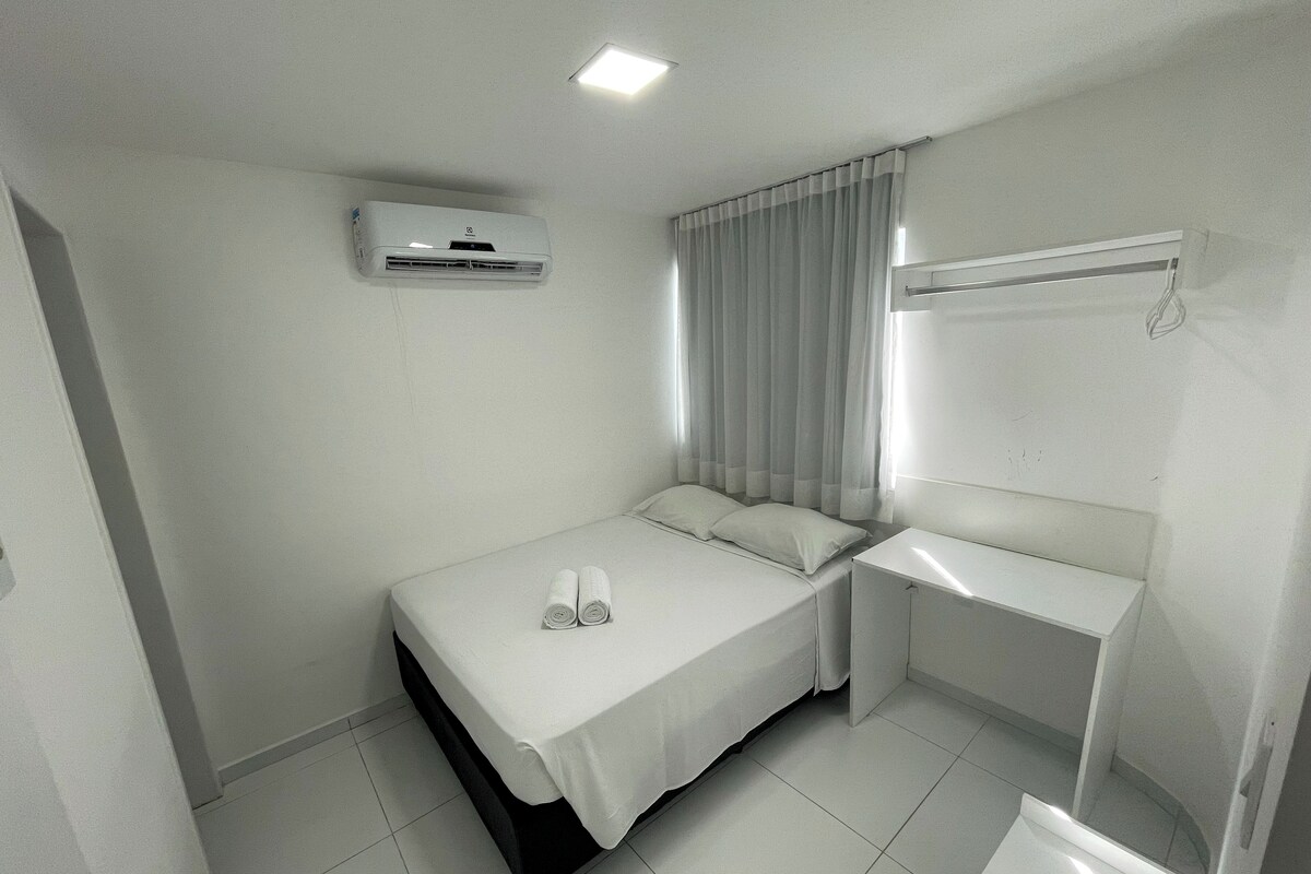 A small, clean room with a bed, a bedside table, a mirror, and an air conditioning unit.